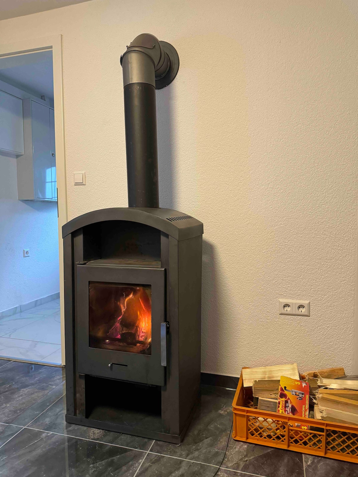 A modern wood-burning stove is positioned against a light, textured wall. The stove has a sleek black finish and glass front, displaying a gentle flame inside. Next to it, a basket holds an assortment of books, complementing the cozy environment.
