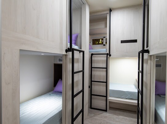 A spacious capsule room features multiple sleeping areas with comfortable beds and soft purple pillows. Each bed is enclosed for privacy, and ladders provide access to the upper levels. Natural light filters in through well-placed openings, creating a serene environment.
