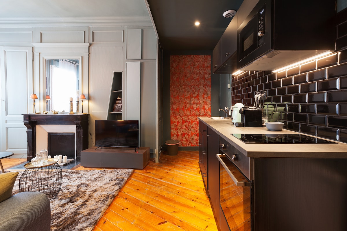 A stylish living area features a comfortable sofa and a flat-screen television. The modern kitchen, equipped with sleek black cabinetry and appliances, is easily accessible. A marble fireplace and decorative accents enhance the room's character, while wooden flooring adds warmth to the space.