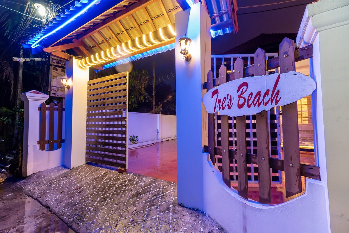 An illuminated entrance gate welcomes guests with vibrant blue lighting. A wooden sign inscribed with 'Tree’s Beach' is prominently displayed, blending with the white walls and ochre tiles of the facade. The pathway is lined with pebbles, adding a natural touch to the approach.