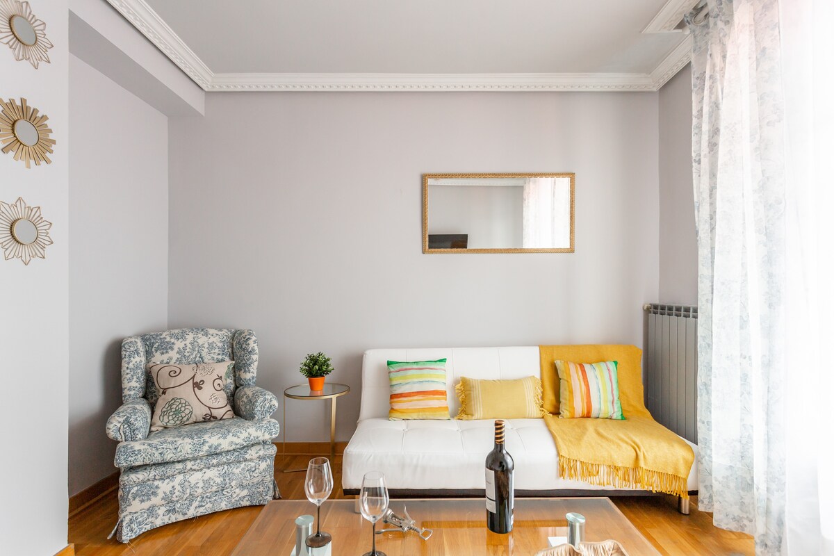 A comfortable living area presents a white sofa adorned with colorful pillows and a bright throw. A floral patterned armchair complements the space. Natural light floods in through sheer curtains, highlighting a small plant and a decorative mirror on the wall.