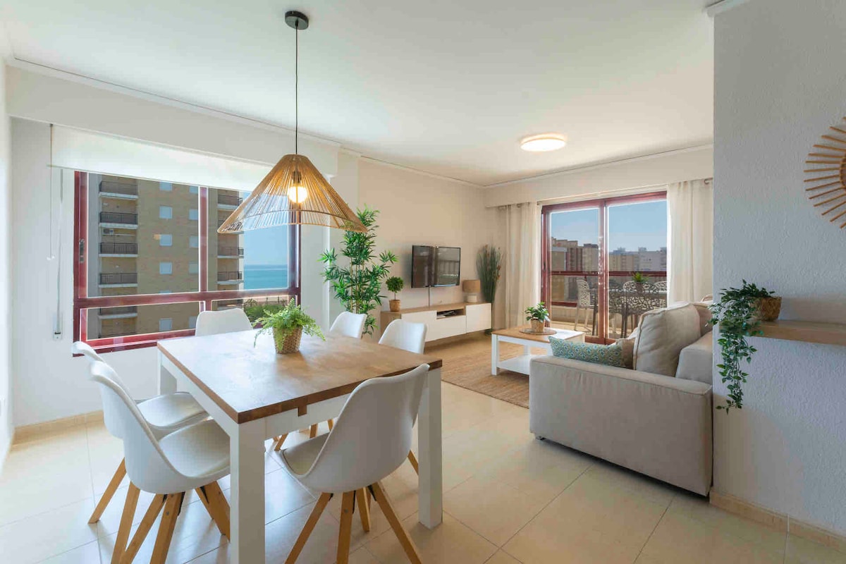 A bright living space features a dining table surrounded by six modern chairs. A comfortable sofa rests nearby, facing a media unit and a large window offering scenic views. Natural light floods the room, enhanced by a stylish pendant light above the table.
