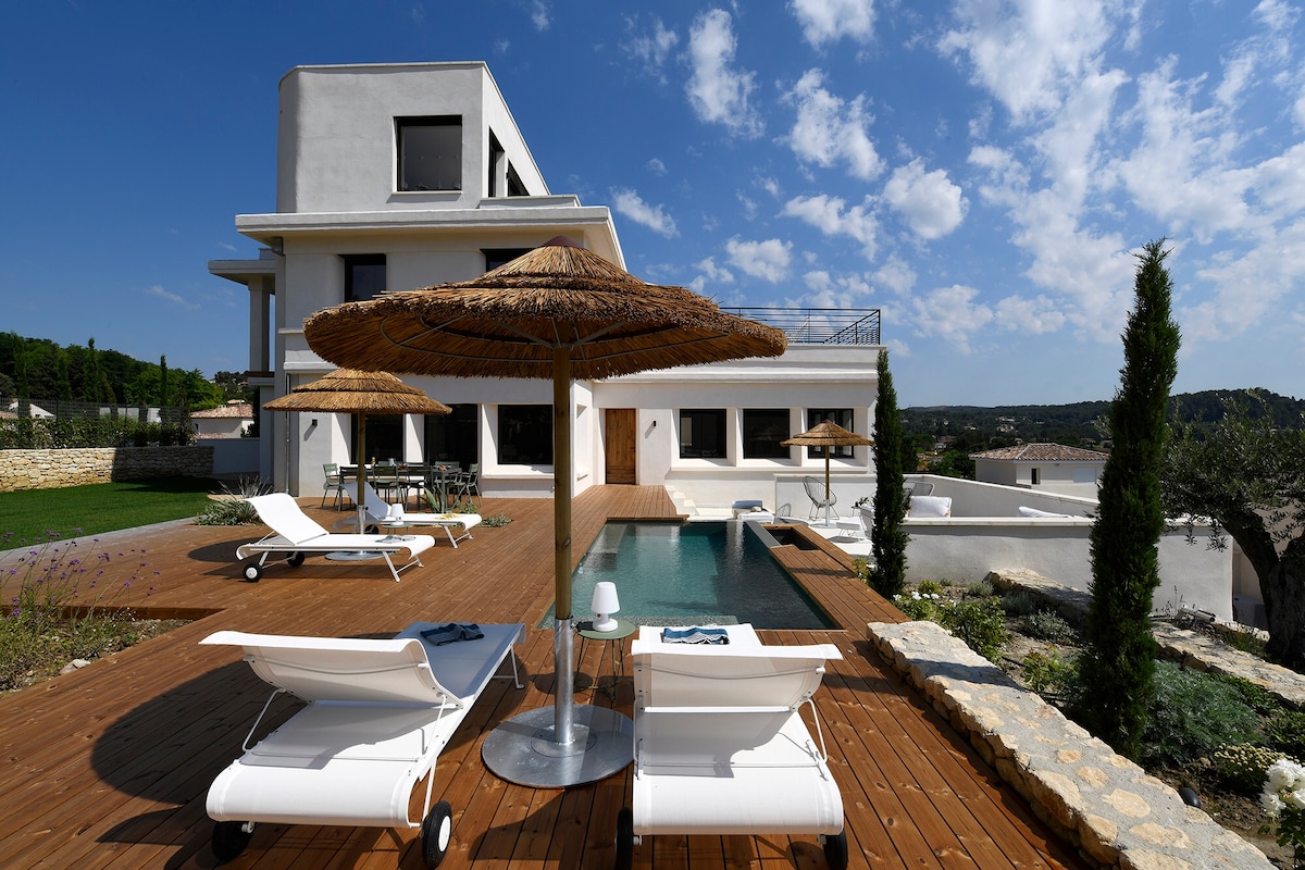 This outdoor space features a wooden deck surrounding a modern infinity pool, complemented by two lounge chairs. A round, straw parasol offers shade, while the backdrop showcases the villa's contemporary architecture against a blue sky dotted with clouds.