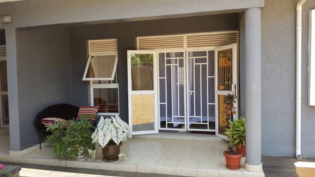 An entrance area showcases a pair of glass doors framed by simple white patterns. A small seating area features a dark sofa adorned with striped cushions. Potted plants add greenery, while tiles provide a clean and inviting floor space.