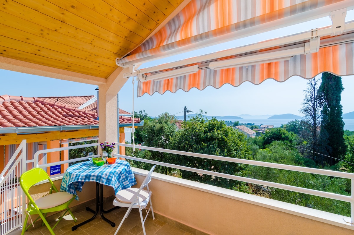 A balcony is shown with a table set for two, featuring a checkered blue and white tablecloth. Colorful folding chairs are positioned around the table. A retractable awning provides shade, and lush greenery and the sea are visible in the background.