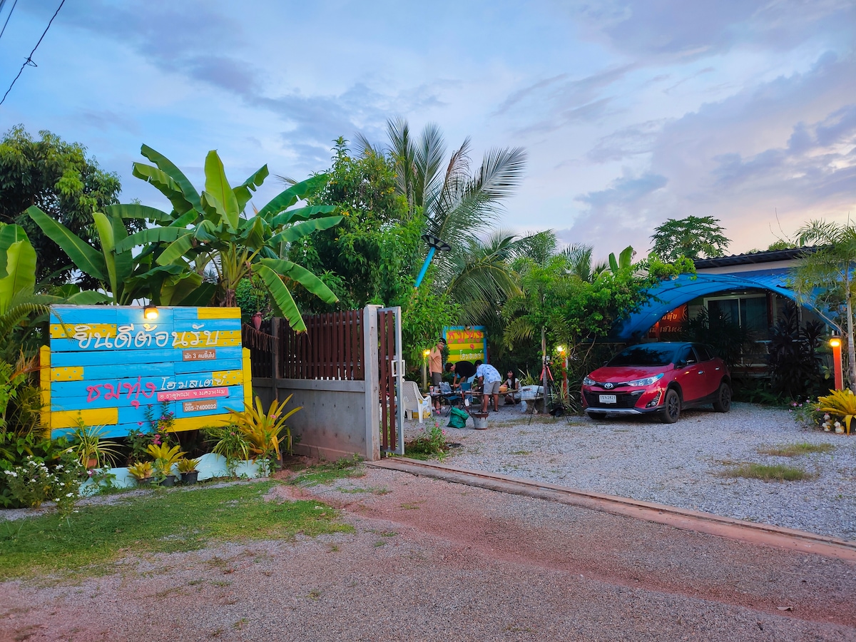 The entrance to the homestay is lined with tropical plants and features colorful signage. A gravel pathway leads to the welcoming area, where guests can be seen enjoying the outdoor space. A red car is parked nearby, under a serene sky filled with soft clouds.