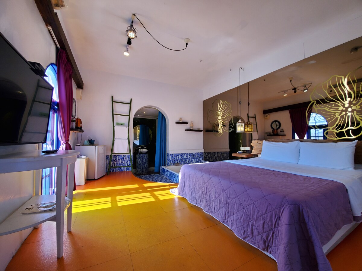A spacious room features a large bed covered with a neat lavender quilt. Natural light filters through colorful curtains onto the vibrant yellow floor. A TV is positioned against one wall, while unique decor elements, including a stylish wall design and a decorative ladder, enhance the atmosphere.