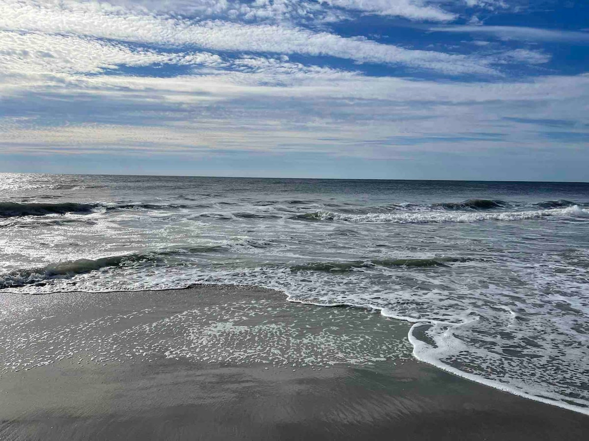 The image captures a serene beach scene, where gentle waves lap against a sandy shore. A vast expanse of ocean stretches into the distance under a sky filled with soft clouds, reflecting hues of blue and gray.