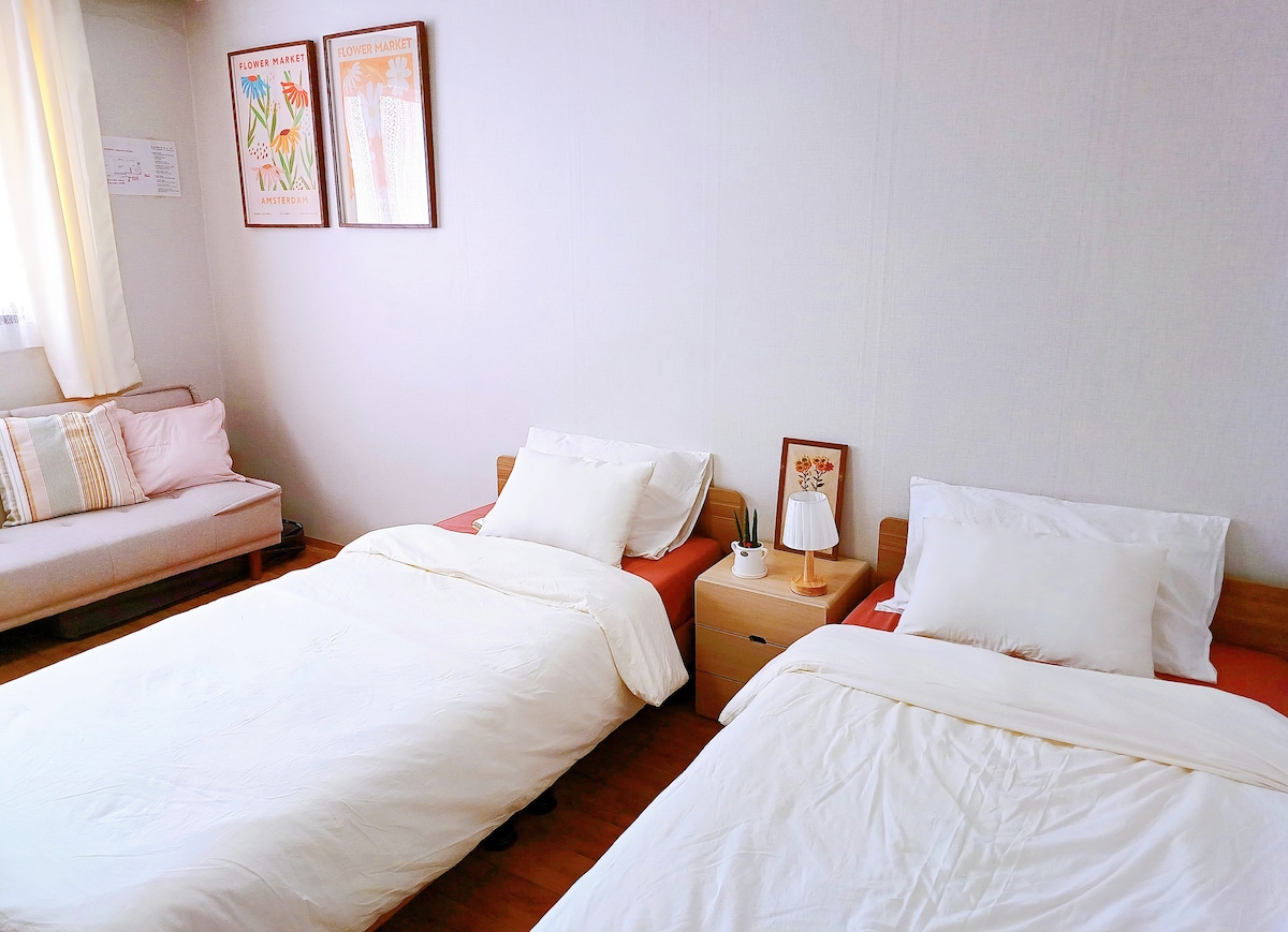 A cozy bedroom features two single beds with fresh white linens. A side table with a lamp is placed between them. A small sofa is situated against the wall, accompanied by soft cushions. Natural light fills the room through a large window adorned with light curtains.