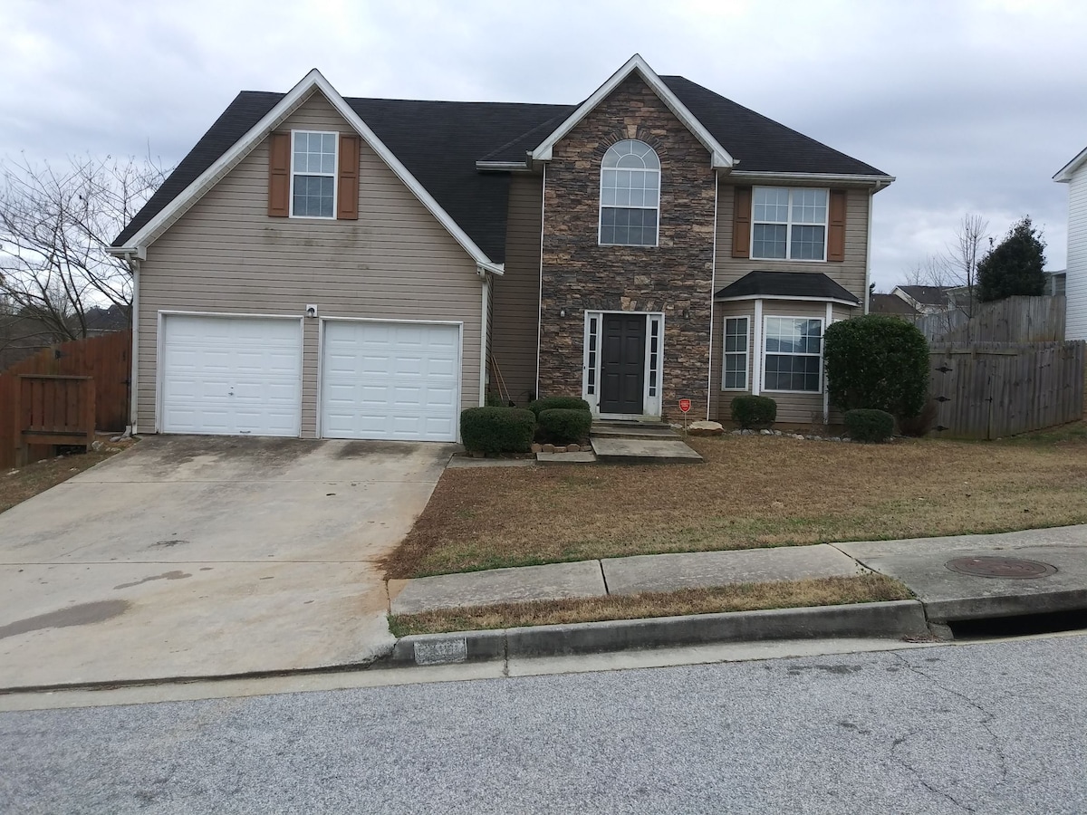 A two-story house is depicted with a combination of beige siding and stone accents. The front features two garage doors and a walkway leading to the entrance. Green shrubs frame the door, while a fenced yard adds a layer of privacy.