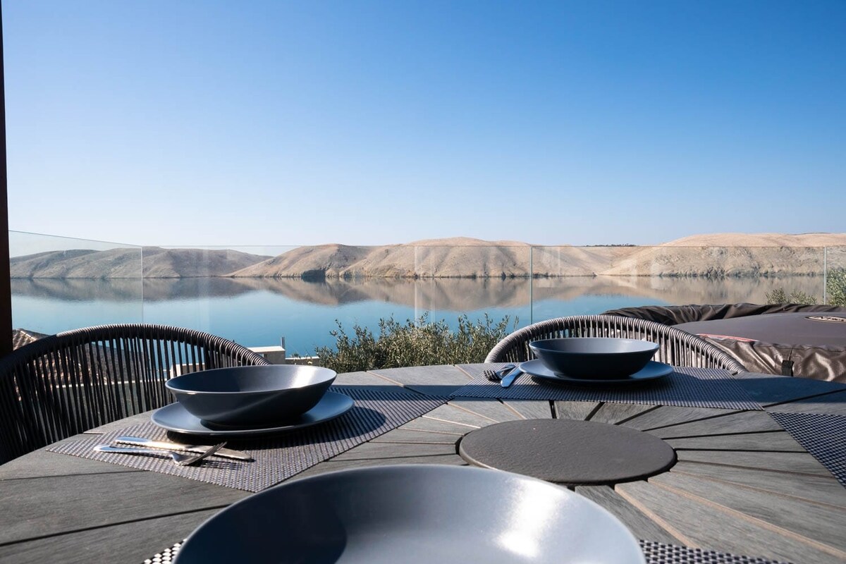 A dining table is set outdoors, featuring dark plates and silver utensils. The view includes shimmering water bordered by sandy hills under a clear blue sky. Nearby, a jacuzzi is partially visible, enhancing the serene atmosphere of the terrace.