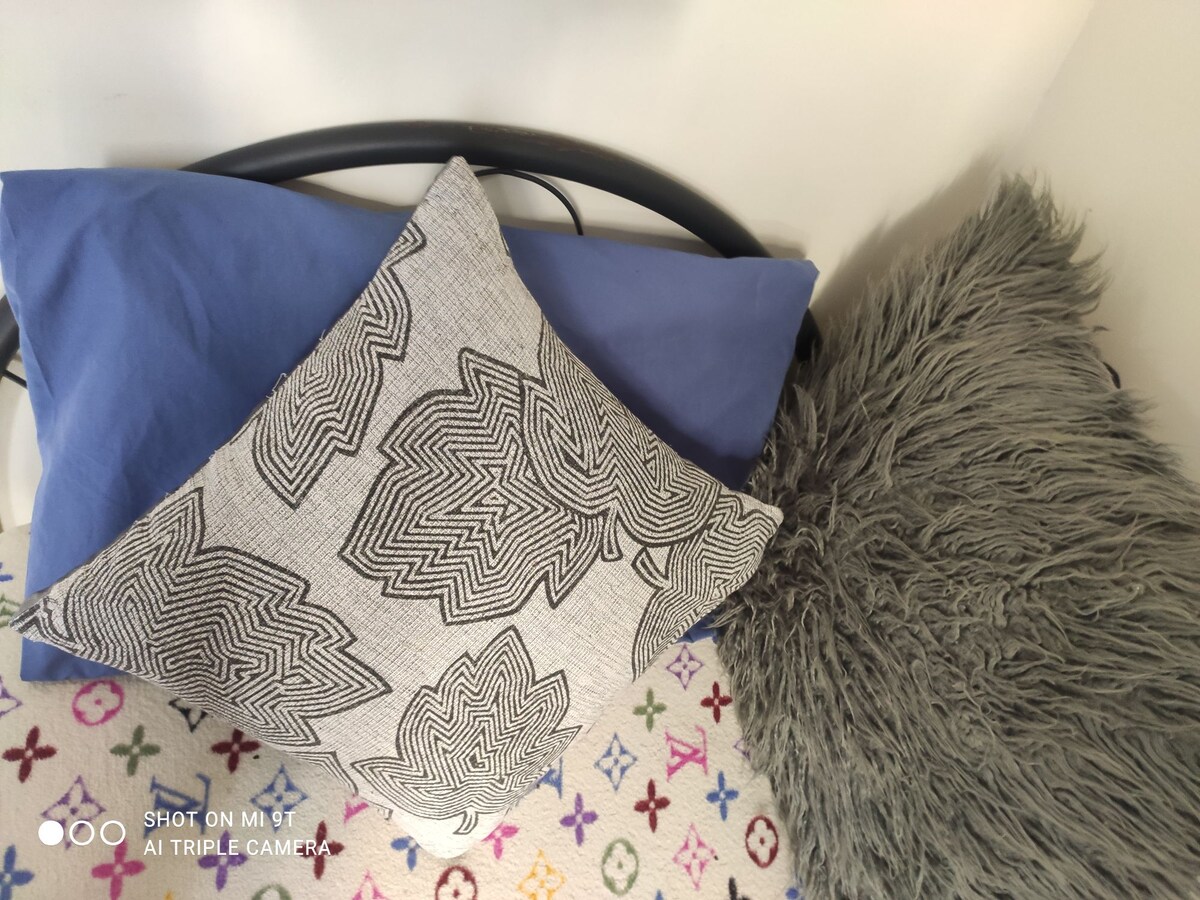 A close-up view of a neatly arranged bed featuring a blue pillow and a textured throw pillow with a patterned design. A fluffy grey cushion adds a touch of comfort, while a colorful embroidered blanket is visible beneath them.