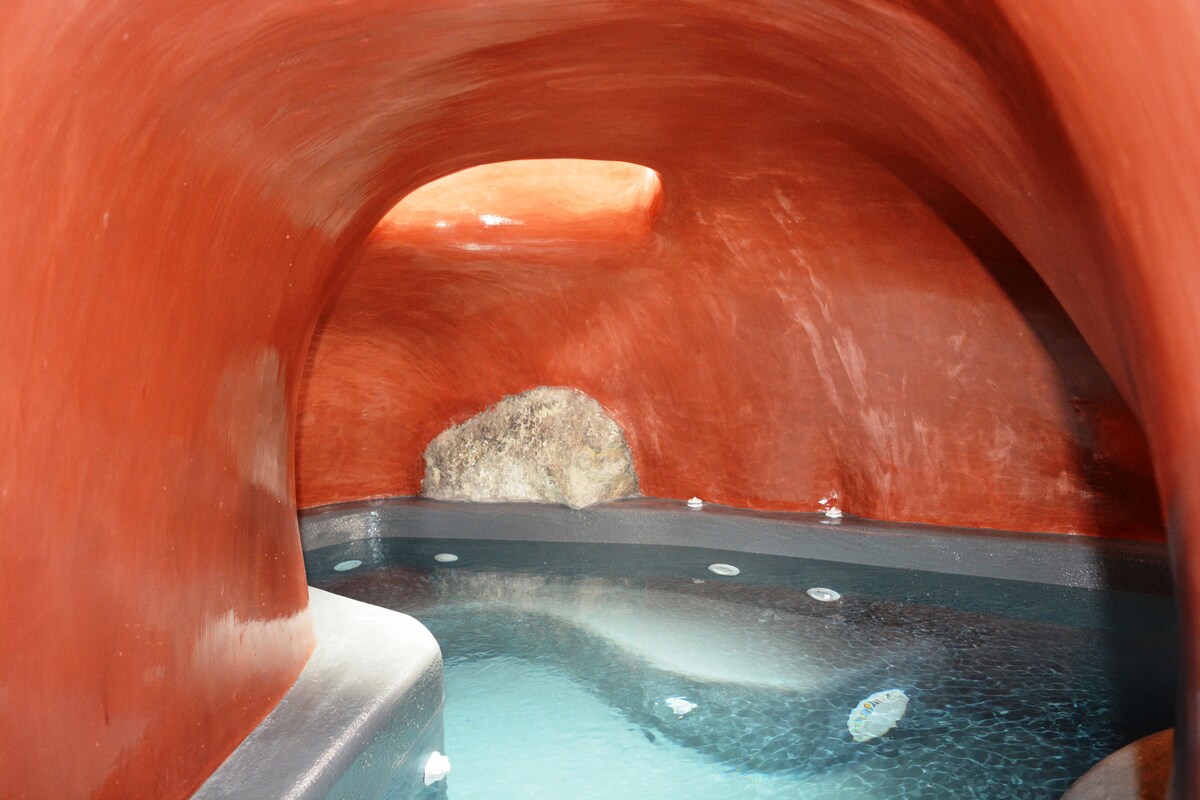 An indoor cave plunge pool is featured within a uniquely shaped cave, surrounded by smooth red mortar walls. Natural rock adds texture to the setting, while the water surface reflects soft light, creating a serene atmosphere.