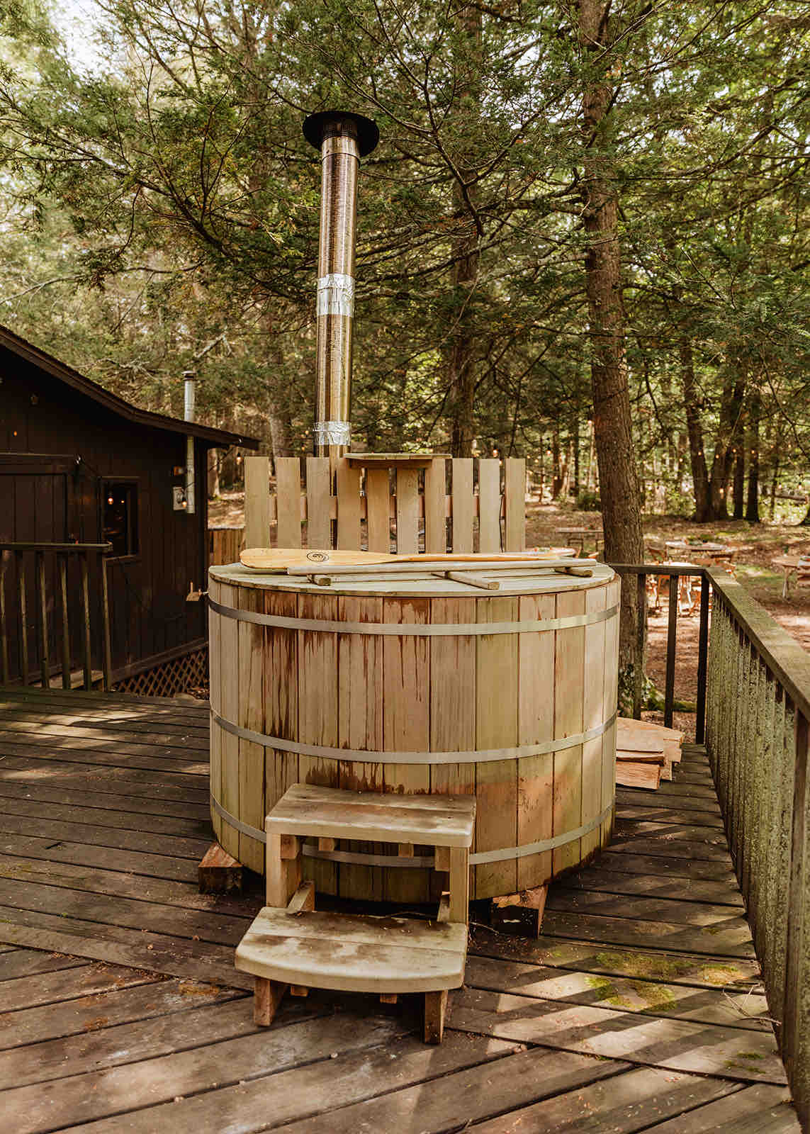 A wood-fired cedar barrel hot tub is situated on a wooden deck, surrounded by tall trees. The hot tub features a chimney for ventilation and steps for easy access, providing a relaxing space in a secluded outdoor setting.