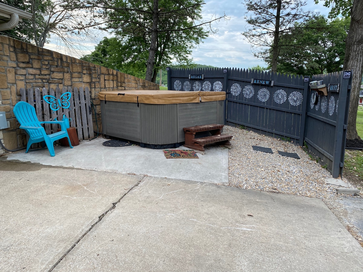 A private outdoor space features a hot tub with a wooden cover, surrounded by a gravel area and bordered by a wooden fence adorned with decorative designs. A light blue chair provides a seating option next to decorative wooden panels.