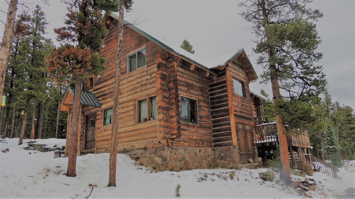A rustic log cabin is nestled among snow-covered ground and pine trees. The structure features multiple windows and a welcoming deck, seamlessly integrated with the natural surroundings. The building's wooden exterior highlights the craftsmanship, while the winter landscape enhances the serene atmosphere.