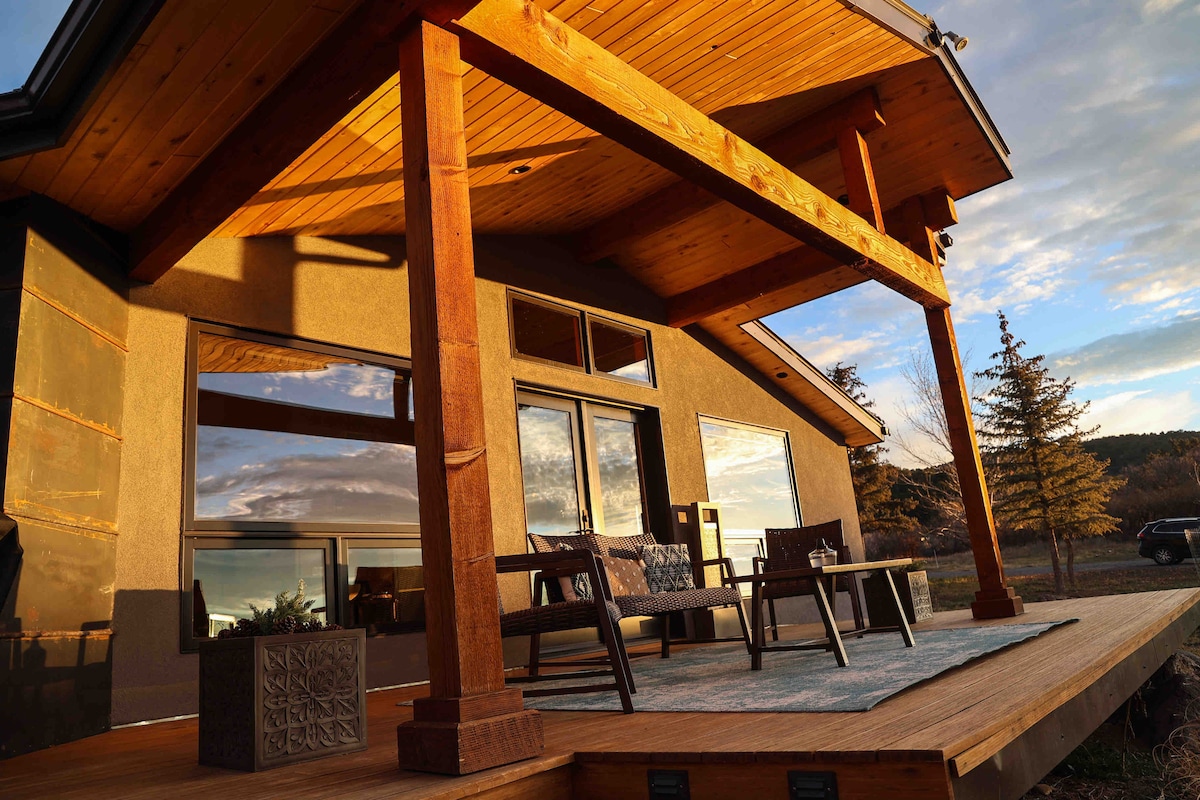 A covered porch is supported by wooden beams, featuring outdoor seating arranged on a textured rug. Large windows reflect the golden hour light, blending the indoor and outdoor spaces. Scenic views extend beyond the deck, highlighting the natural surroundings.