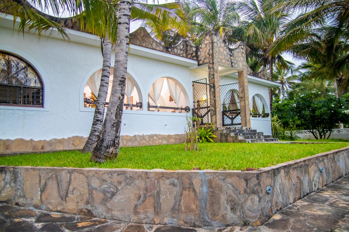 The exterior of the home features a white façade framed by tall palm trees. Stone steps lead to a decorative entrance with archways, complemented by a grassy area in front and a natural stone wall, creating a serene and welcoming environment.