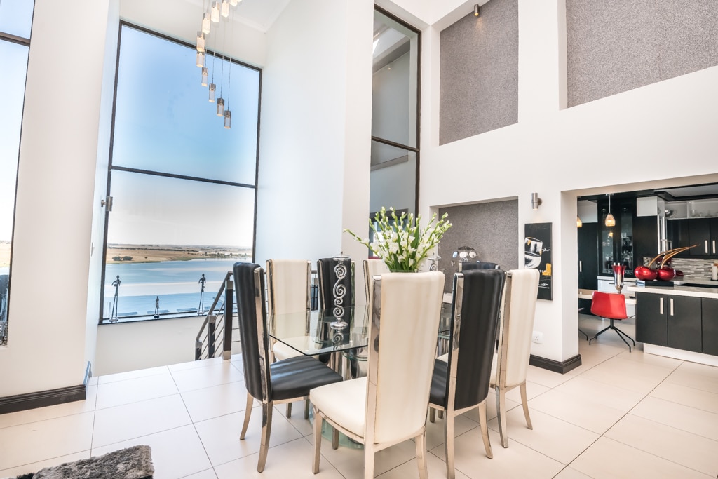 A modern dining area with a glass table is framed by six high-backed chairs in alternating colors. Large windows provide a view of the water, allowing natural light to fill the space. A stylish chandelier adds an artistic touch above the table.