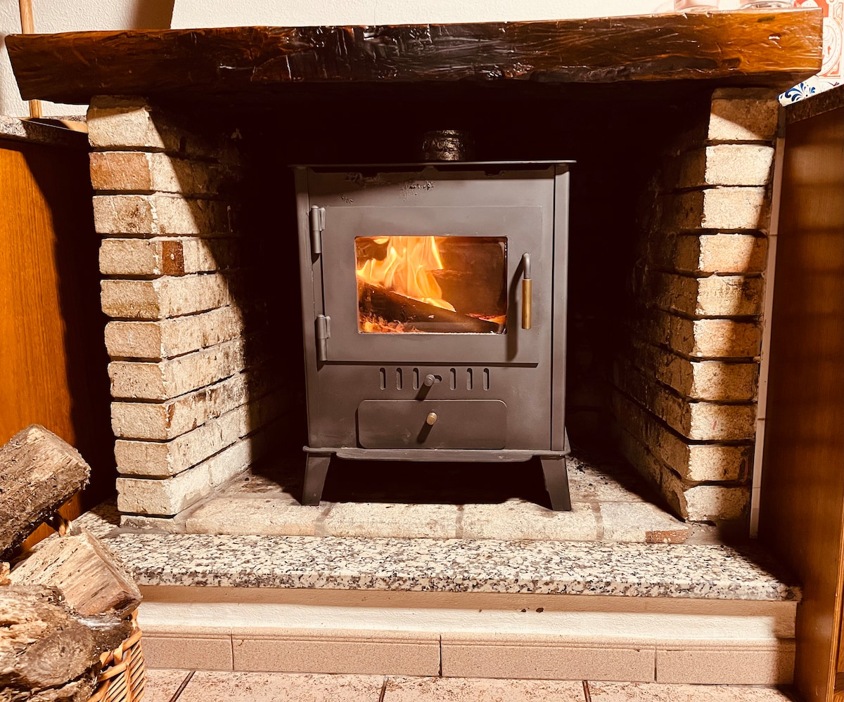 A wood-burning stove is positioned in a brick alcove with a stone base, featuring visible flames inside. Logs are stacked beside the stove, and the surrounding area is complemented by warm tones of wood and brick, enhancing the cozy atmosphere.