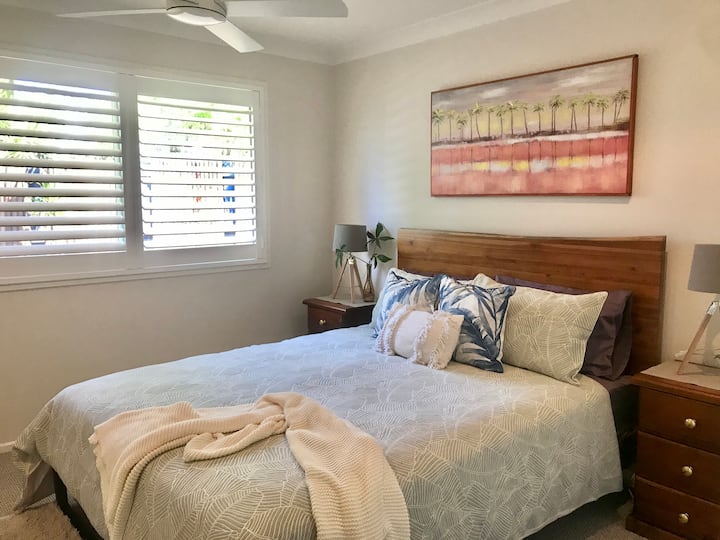 A cozy bedroom with a wooden bed frame, soft bedding in neutral tones, and decorative pillows. Two wooden bedside tables with lamps flank the bed. A large window with plantation shutters lets in natural light, and a framed painting of palm trees hangs above the bed