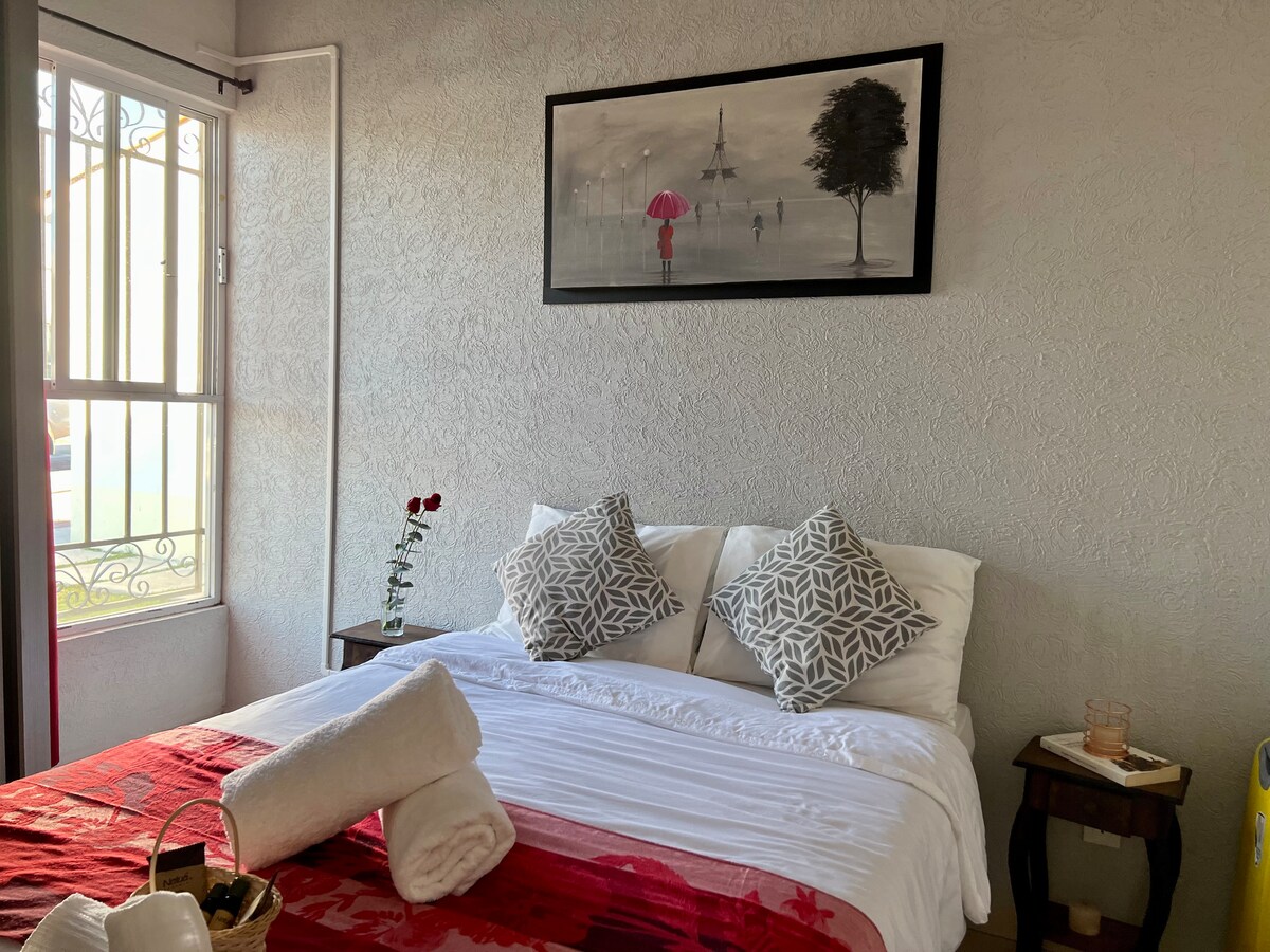 A bright and inviting bedroom features a spacious bed with white linens and decorative pillows. A red and white blanket adds a touch of color. A small table beside the bed is adorned with a rose, while natural light filters through a window, enhancing the serene atmosphere.