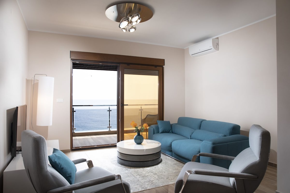 A spacious living area features a light blue sofa and two armchairs, arranged around a circular coffee table. A large balcony door offers a view of the sea, allowing natural light to fill the space. A flat-screen TV is mounted on the wall.