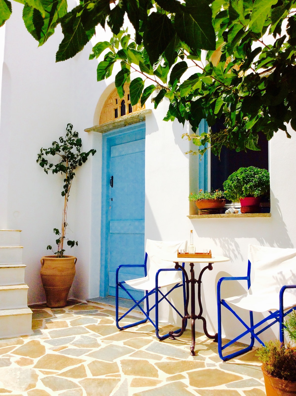 A charming outdoor seating area features two white chairs on a stone-paved patio, accompanied by a small table. A potted plant sits nearby, and a vibrant blue door with a wooden top is visible, framed by white walls and greenery overhead.
