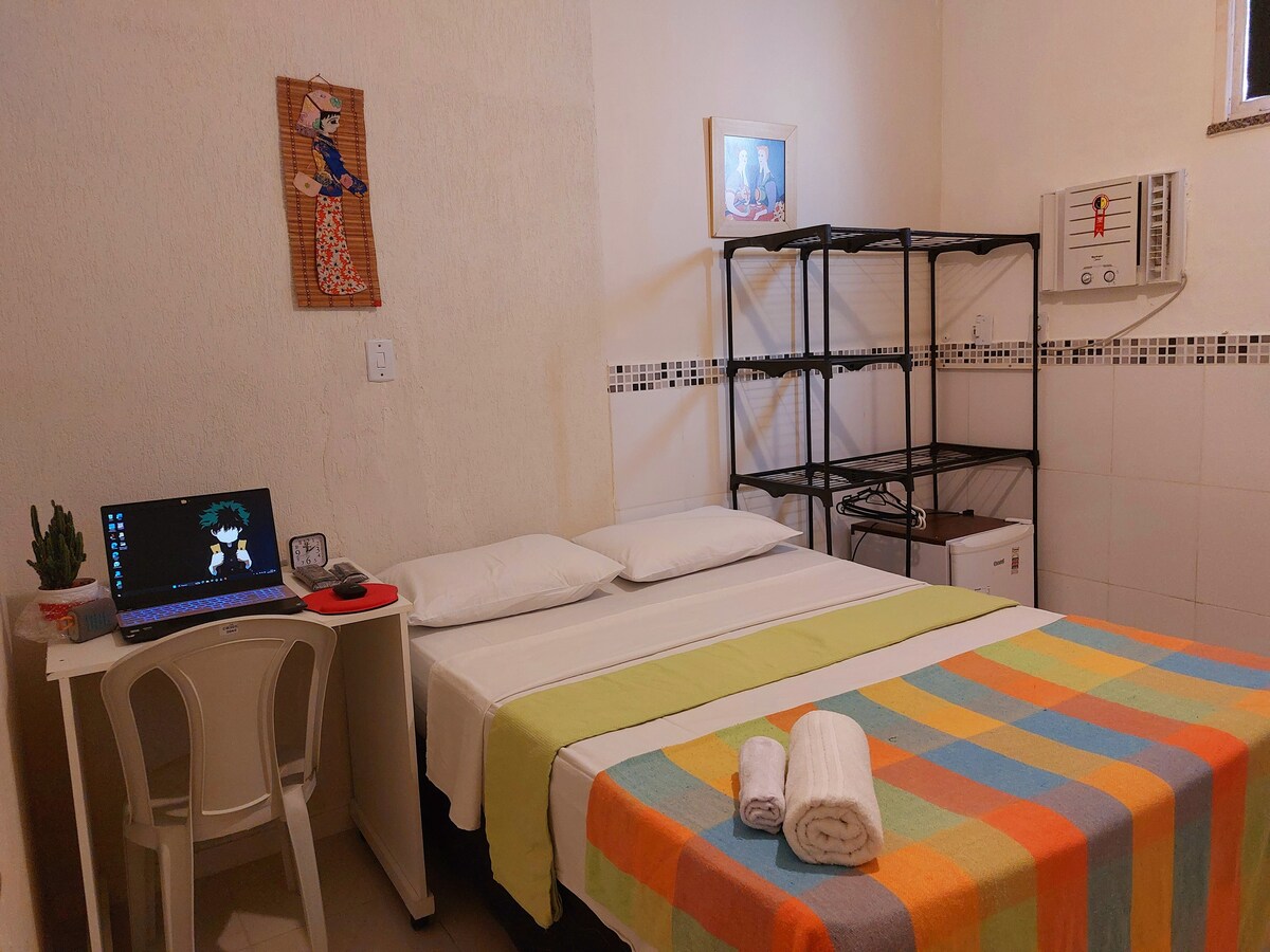 A cozy room features a comfortable bed with a colorful blanket, complemented by white linens. A desk with a laptop sits beside a potted plant. Shelving units and a wall-mounted air conditioning unit are visible, offering convenient storage and climate control.