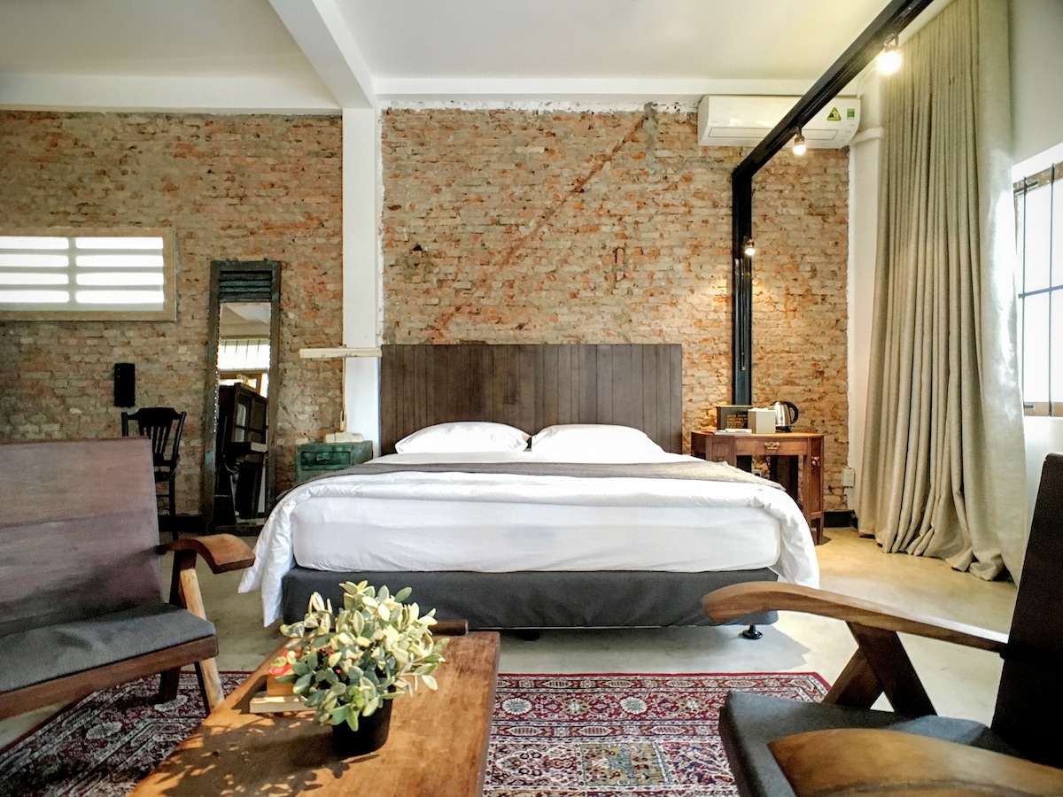 The spacious loft features exposed brick walls and a stylish wooden bed frame supporting a comfortable mattress. Soft curtains frame a window, and a cozy seating area is arranged with modern chairs around a rustic wood coffee table. A kettle is placed on a side table, adding convenience.