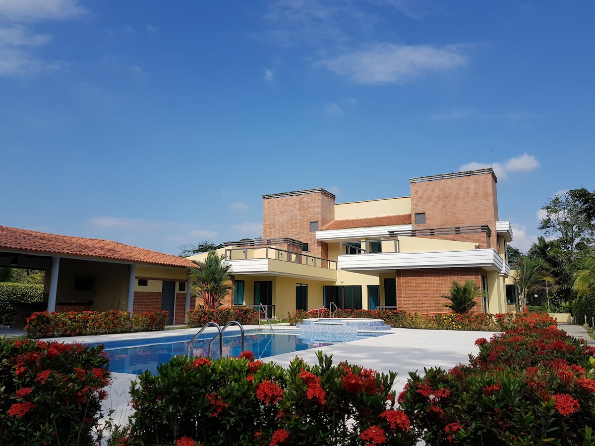 A spacious house with a multi-level design is surrounded by vibrant flower bushes. A clear swimming pool is visible in front of the house, reflecting the blue sky. Lush greenery and palm trees add to the natural setting, creating a tranquil atmosphere.