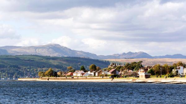 A scenic view shows a waterfront lined with a variety of houses and trees, framed by distant mountains. The calm water reflects the soft clouds overhead, contributing to a serene atmosphere.