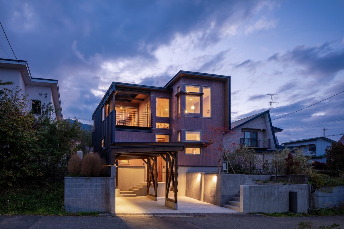 The two-story house features a modern wooden exterior with large windows that reflect the evening sky. Soft lighting highlights the entryway and outdoor staircase, creating an inviting appearance as dusk settles around the property.