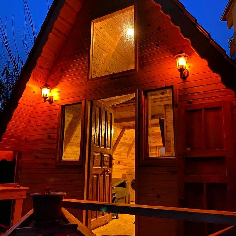 A charming wooden bungalow is illuminated by warm outdoor lighting, highlighting the natural wood finishes. The double doors, framed by two wall lamps, open into a cozy interior space that features wood-paneled walls and a welcoming ambiance.