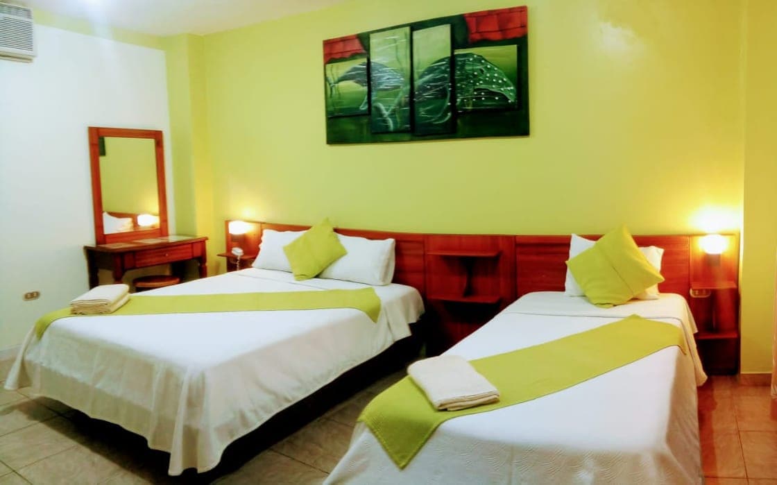 A bright and airy guest room features two spacious beds, each adorned with green accents. A mirror and a wooden table are positioned in the corner. Soft lighting from bedside lamps creates a warm ambiance, while a vibrant artwork of leaves decorates the wall above the beds.