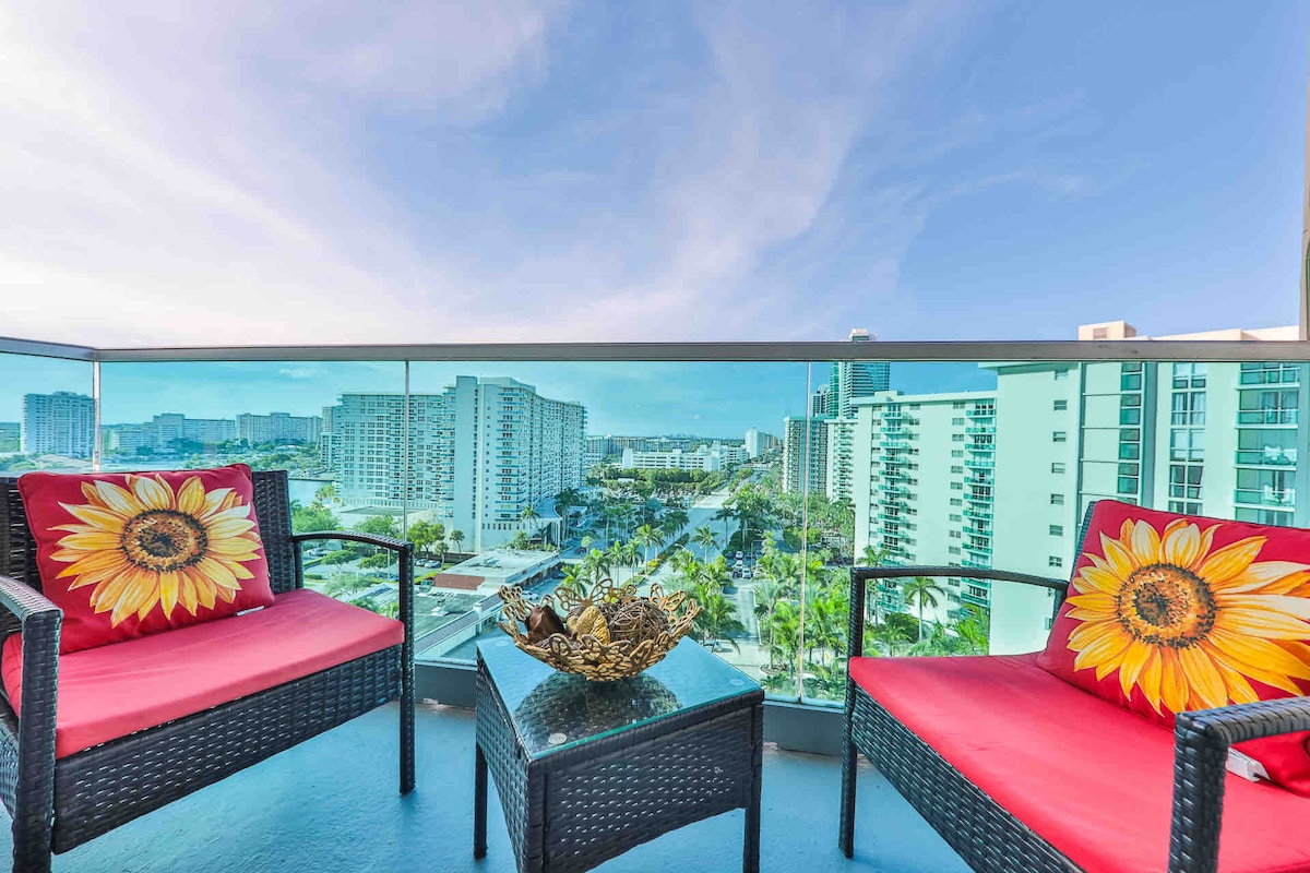 A balcony view features two wicker chairs adorned with sunflower cushions, offering a bright and cheerful spot to relax. A small table with a decorative bowl sits in between, while expansive city and ocean views stretch into the horizon, under a clear blue sky.