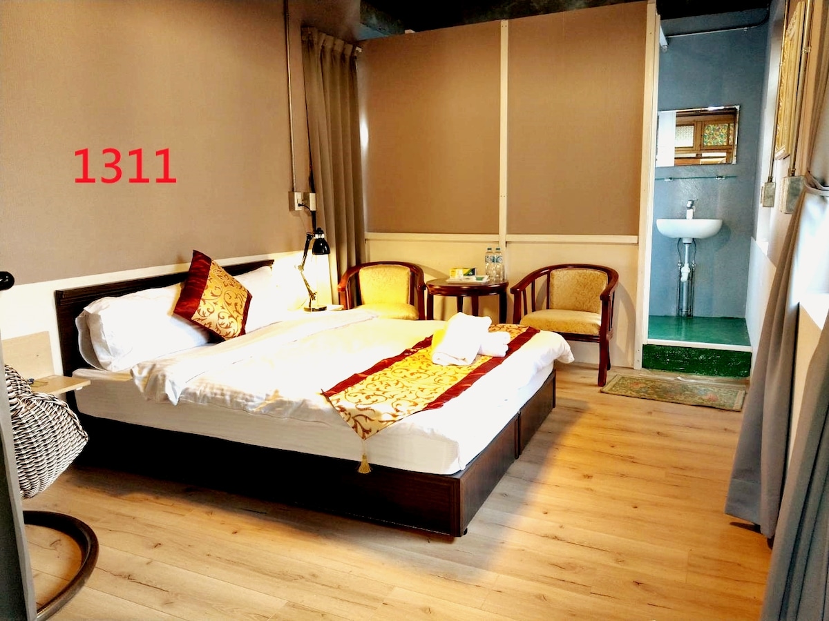 A cozy room features a spacious bed adorned with decorative pillows. Two armchairs are positioned beside a small table, ideal for relaxation. A private bathroom is displayed in the background, providing convenience and privacy.