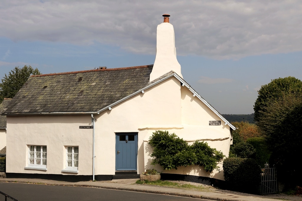 The cottage exterior features a whitewashed facade complemented by a blue front door. A prominent chimney rises from the roof, and trimmed shrubs frame the entrance, enhancing the quaint appearance of this charming period home.