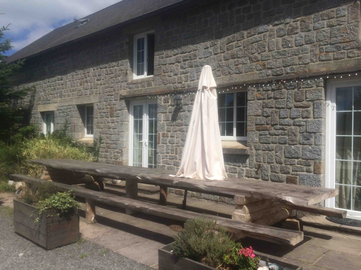 An outdoor dining area features a large wooden table, covered with a protective cloth. Surrounding planters with greenery enhance the space, while stone walls of the gite provide a rustic backdrop. Sliding glass doors are visible, leading into the indoor area.