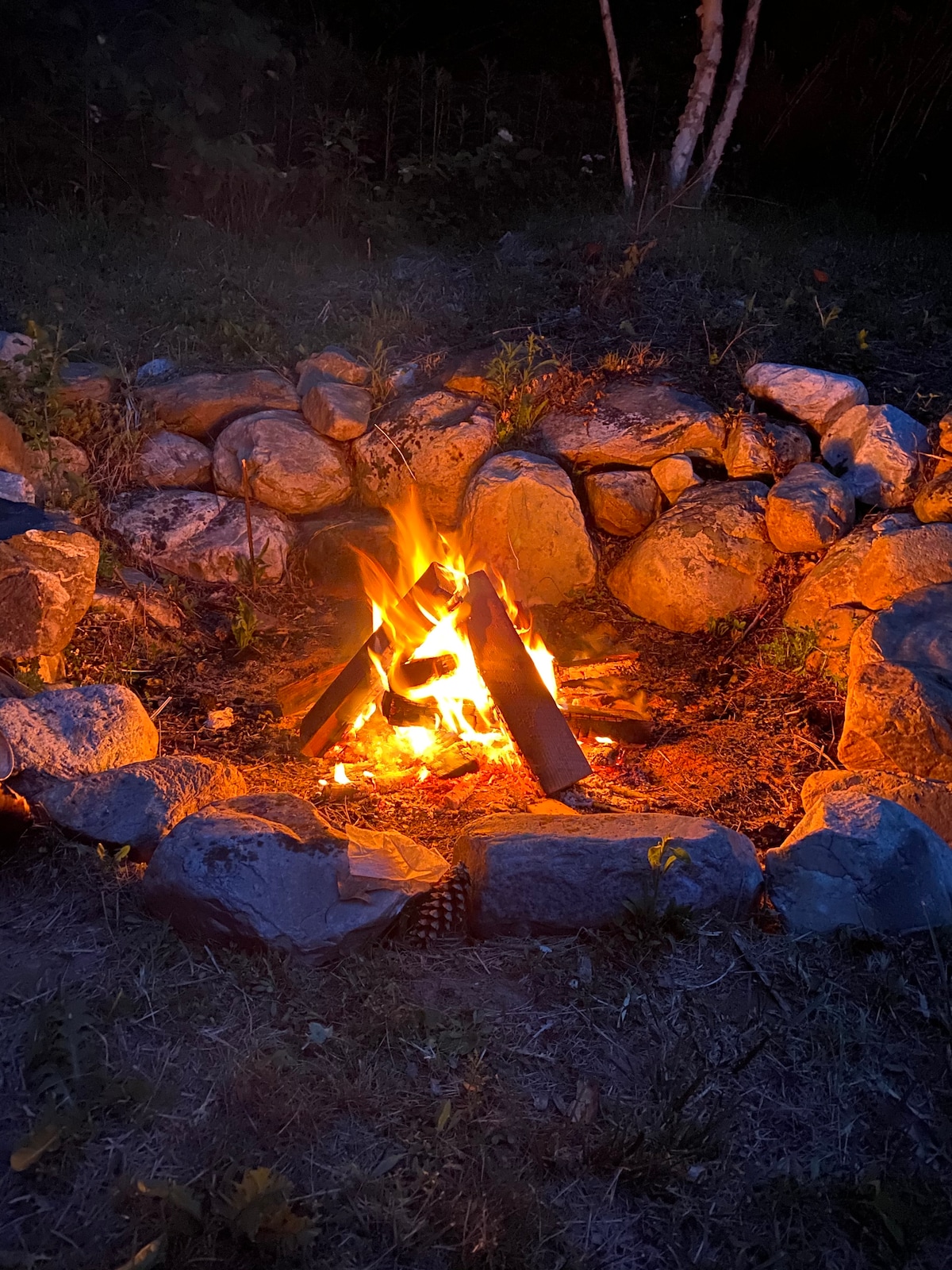 A cozy fire pit is surrounded by a circular arrangement of stones. Flickering flames rise from the center, illuminating the nearby rocks and providing a warm glow to the evening setting. Dark surroundings contrast with the bright fire, creating a peaceful outdoor ambiance.