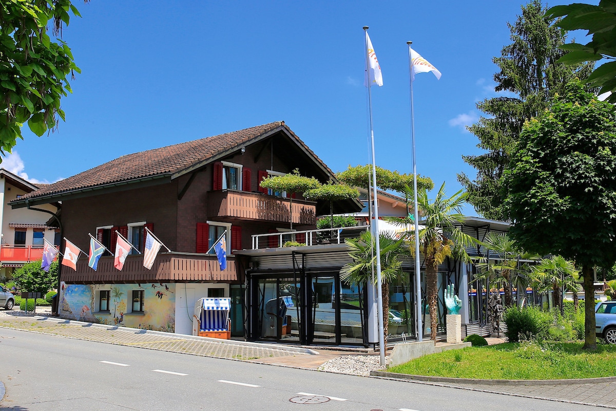 The chalet-style building, showcasing a combination of traditional and modern architecture, is surrounded by greenery. Several flags are displayed in front of the structure, which features large glass windows allowing natural light to enhance the interior.