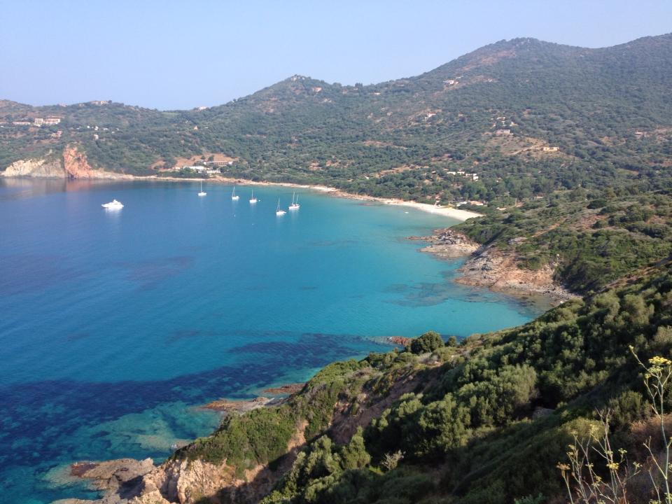 A serene coastline is visible, featuring clear turquoise waters gently lapping against the shore. Several boats are anchored in the bay, with verdant hills rising in the background, showcasing a blend of green foliage and rocky terrain.