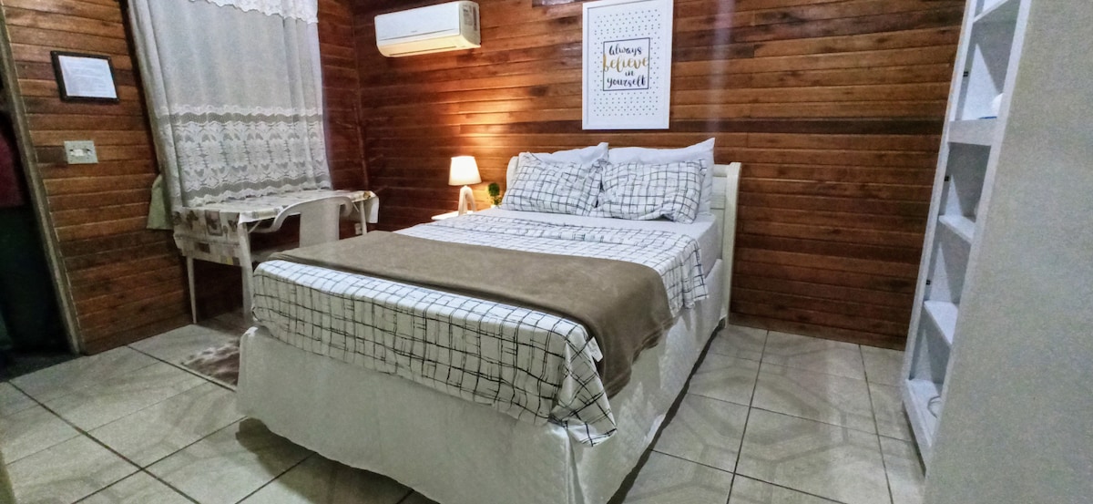 A cozy sleeping area is presented with a spacious bed dressed in checked linens. Wooden paneling creates warmth, while a small lamp provides soft lighting. An air conditioning unit is mounted above, and a decorative wall piece complements the room's ambiance.