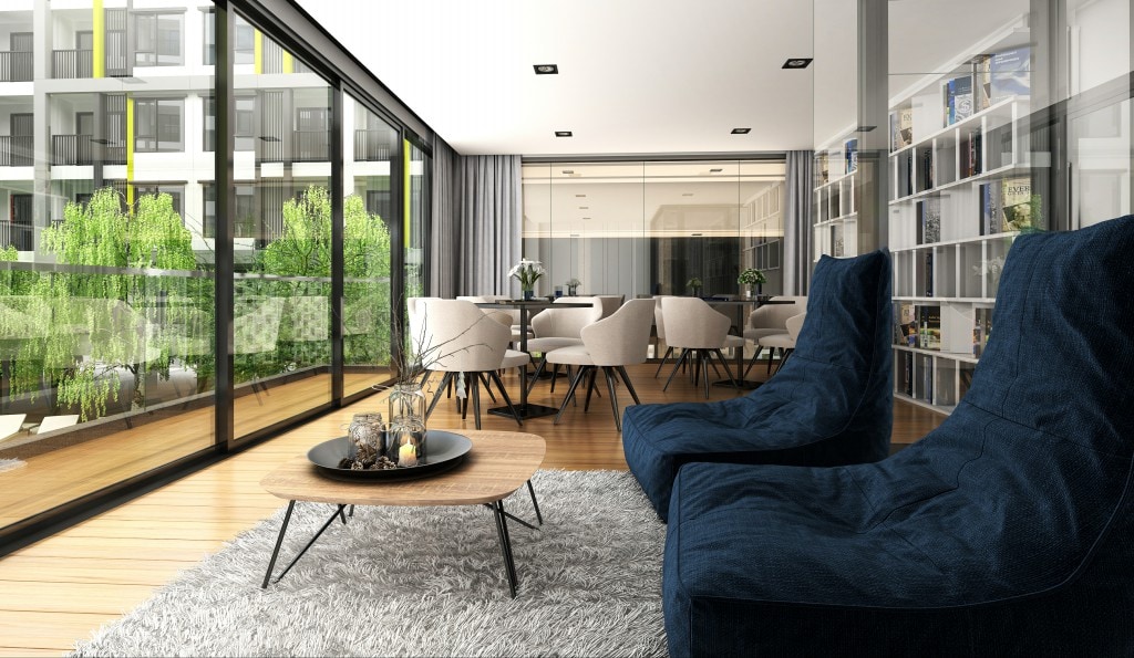 A spacious living area features large glass doors that open to a balcony, allowing natural light to fill the room. Comfortable seating is paired with a circular coffee table, and a soft area rug enhances the modern decor. Greenery is visible outside, adding a touch of nature.