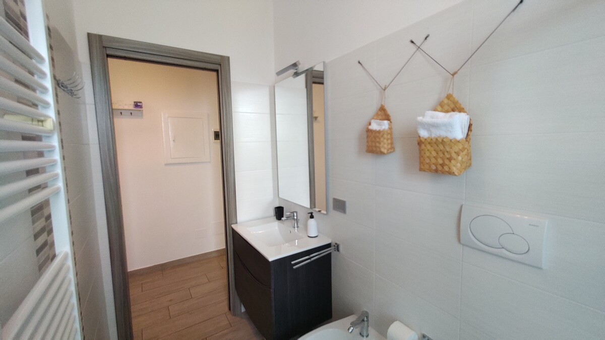 A modern bathroom is depicted, featuring a sink with a dark cabinet and a large mirror. Shelving holds neatly arranged towels in woven baskets. A bathtub is situated next to a stylish radiator, with light-colored tiles covering the walls and floor.