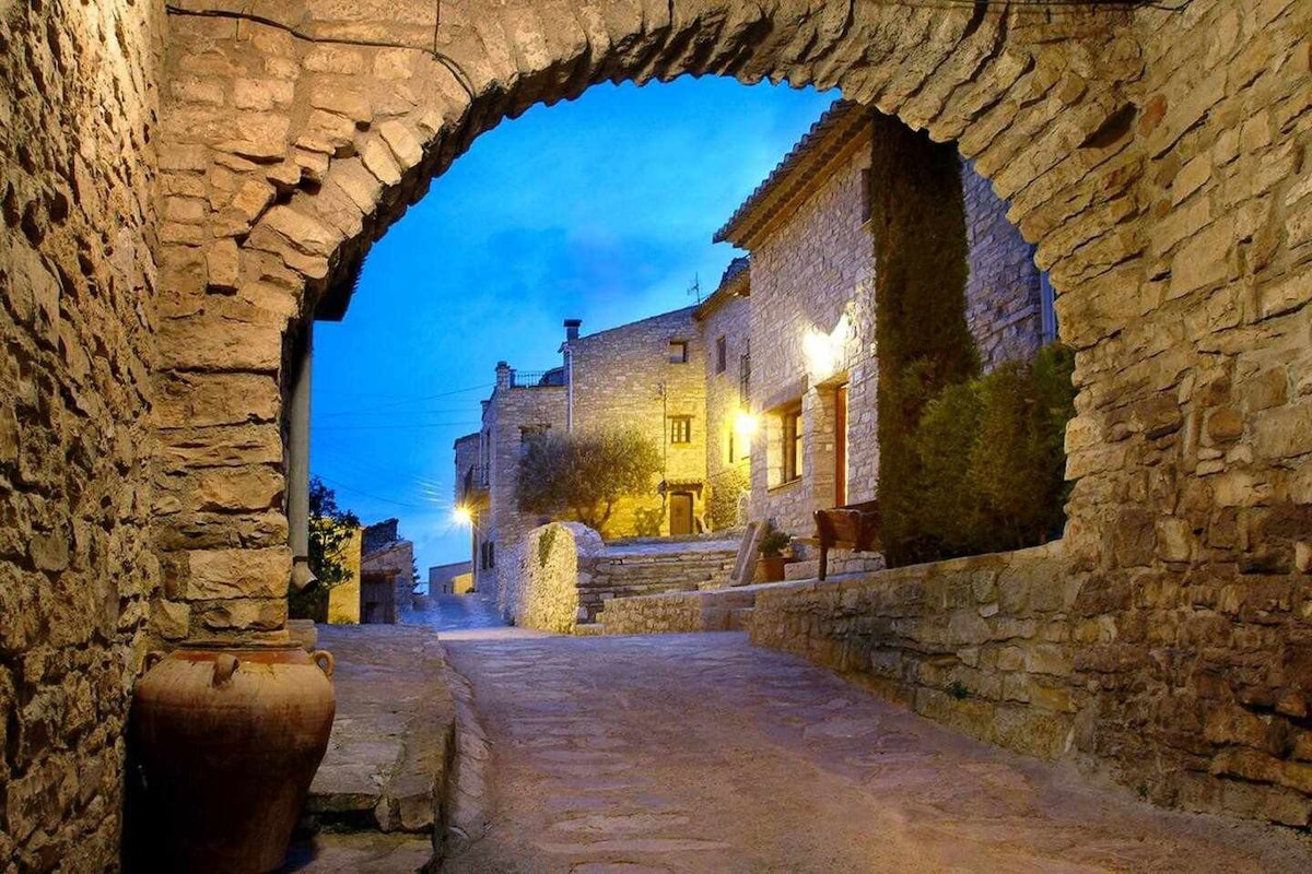 The image depicts a cobblestone path leading through an arched stone passageway. Soft evening light illuminates the surrounding historic buildings, showcasing a blend of rustic stone textures. A large terracotta pot rests on the ground, adding to the charming ambiance of the medieval setting.