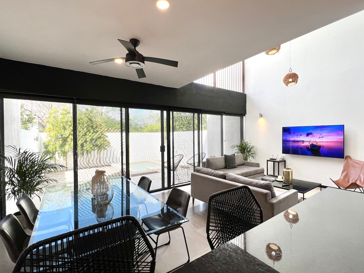 A modern, open-concept living area features large glass doors that enhance natural light. Comfortable seating is arranged around a sleek coffee table, alongside a dining area with a polished table and designer chairs. A wall-mounted television is visible, creating an inviting space for relaxation.