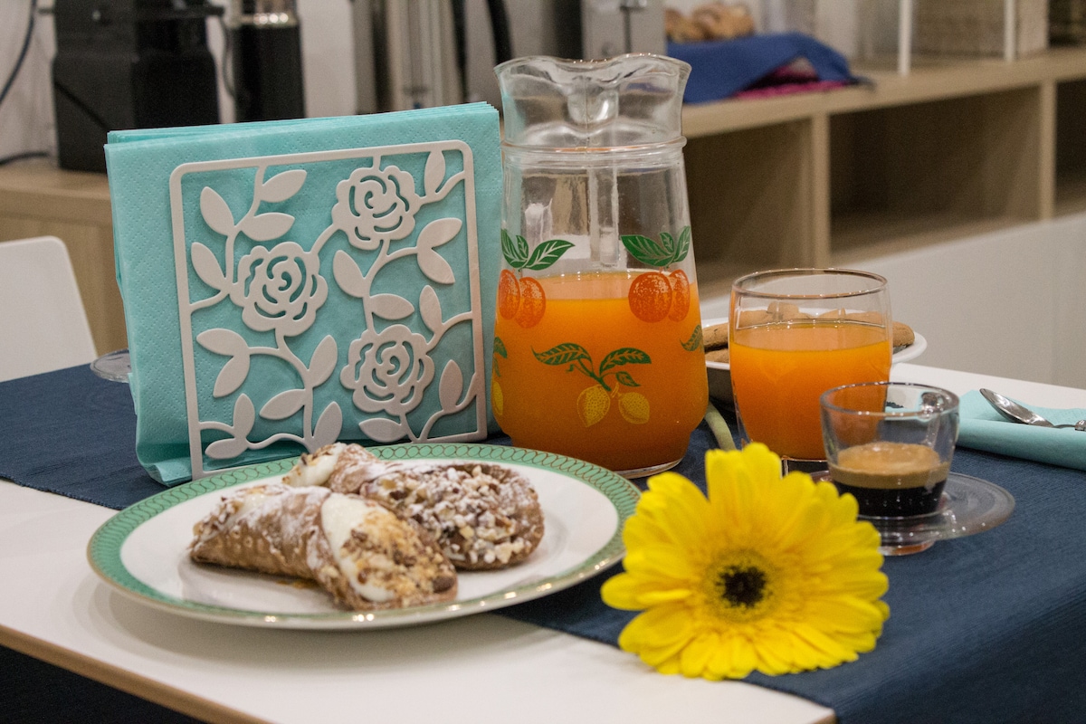 A breakfast setting is presented, featuring a brightly colored pitcher with orange illustrations, a glass of orange juice, and a shot of espresso. Freshly made cannoli are placed on a decorative plate, accented with a sunflower and a blue napkin on a white table.