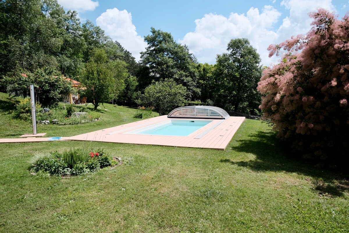An outdoor swimming pool, surrounded by lush green grass and vibrant landscaping, is complemented by mature trees and flowering shrubs. The pool features a covered area, and the tranquil setting invites relaxation and outdoor enjoyment.