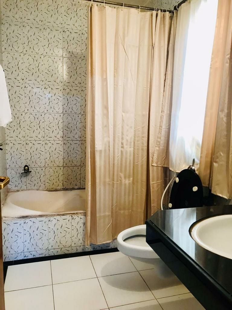 A compact bathroom is designed with light-colored tiles and features a bathtub paired with a shower curtain. A modern sink with a dark countertop is visible beside a toilet. Natural light filters through a window covered by sheer curtains.